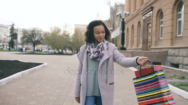 Dolly disparo de Atractiva chica de raza mixta caminando hacia con bolsas de compras y sonriendo a la cámara. Feliz joven mujer caminando después de visitar las ventas del centro comercial — Vídeos de Stock