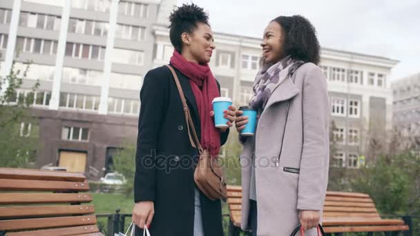 Duas mulheres de raça mista atraentes com sacos de compras bebendo café e conversando na rua. Meninas jovens sorrindo e ter amigos conversa — Vídeo de Stock