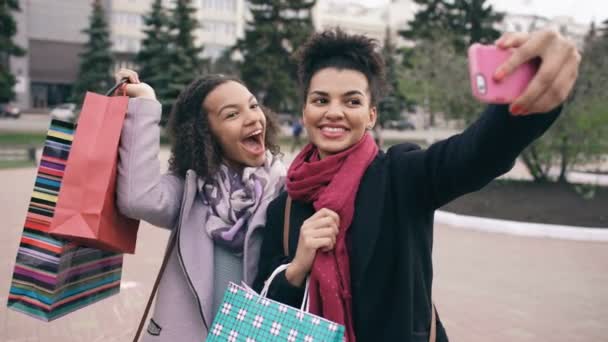 Dos mujeres afroamericanas lindas tomando selfie con bolsas de compras y sonriendo. Amigos se divierten después de visitar la venta del centro comercial . — Vídeo de stock