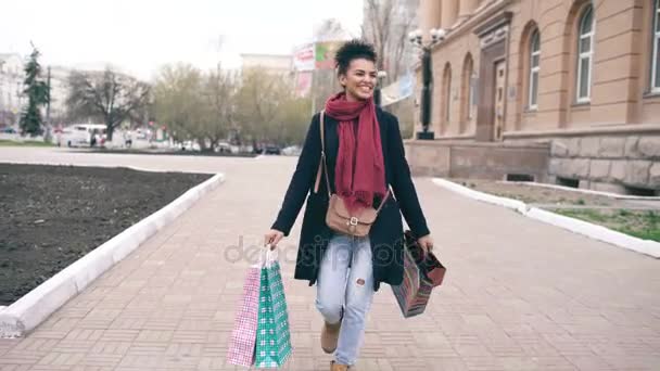 Dolly shot of Attractive mixed race girl dancing and have fun while walking down the street with bags. Happy young woman walking after shopping on mall sale — Stock Video