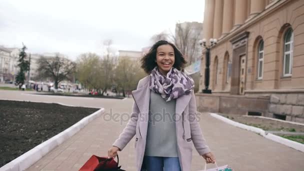 Долли сняла девочку смешанной расы, танцующую и веселящуюся, идя по улице с сумками. Счастливая молодая женщина, гуляющая после шопинга на распродаже — стоковое видео