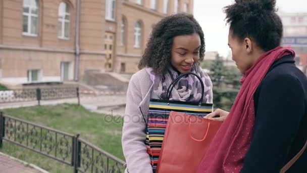 Две молодые африканские женщины делятся своими новыми покупками в пакетах друг с другом. Привлекательные девушки разговаривают и улыбаются после посещения торговых центров продаж — стоковое видео