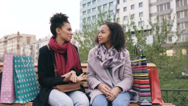 Twee leuke Afro-Amerikaanse vrouw vrienden met boodschappentassen praten na een bezoek aan winkelcentrum verkoop en zitten op bankje — Stockvideo