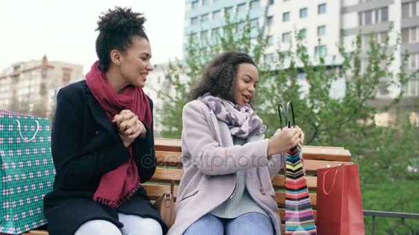 Dos jóvenes afroamericanas compartiendo sus nuevas compras en bolsas de compras entre ellas. Chicas atractivas hablando mientras están sentadas en el banco steet — Vídeos de Stock