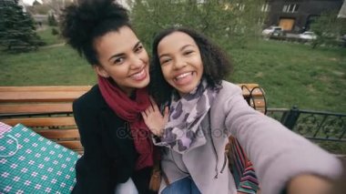Akıllı alışveriş torbaları ile selfie alarak ve gülümseyen iki şirin Afrikalı-Amerikalı kadın. Alışveriş Merkezi satış ziyaret ettikten sonra eğlenceli sokak bankta oturan kız var