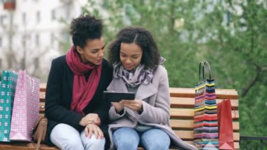 İki çekici karışık yarış kadınla poşetler bankta oturan ve tablet bilgisayar internet sörf için kullanıyor. Mutlu arkadaş alışveriş merkezi satış ziyaret ettikten sonra iyi eğlenceler