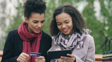 Bankta oturan ve online alışveriş için tablet bilgisayar ve kredi kartı kullanarak kağıt torba ile iki çekici karışık yarış bayanlar