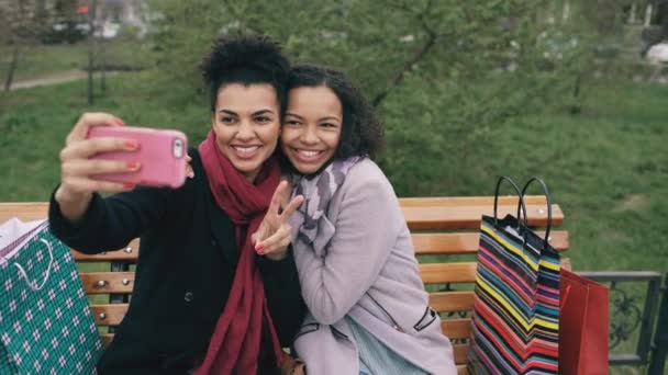 Dwa cute african american kobieta biorąc selfie na smartphone z torby na zakupy i uśmiecha się. Dziewczyny, siedząc na ławce ulicy miłej zabawy po wizycie w centrum sprzedaży — Wideo stockowe