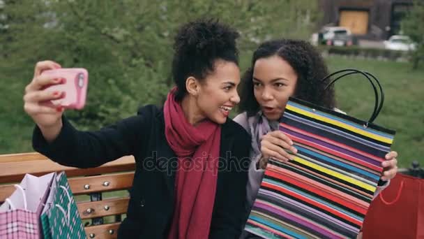 Дві веселі змішані гонки жінки з сумками розмовляють під час відеодзвінків зі смартфоном. Молоді дівчата, які сидять на лавці, спілкуються онлайн з друзями — стокове відео