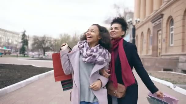 Dolly shot di due attraenti donne di razza mista che ballano e si divertono mentre camminano per strada con le borse della spesa. Felice giovani amici a piedi dopo aver visitato la vendita centro commerciale — Video Stock