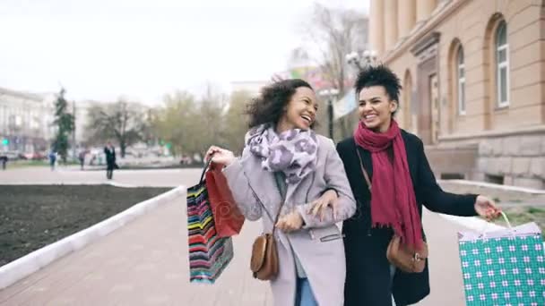 Dolly shot of two attractive mixed race women dancing and have fun while walking down the street with shopping bags. Happy young friends walk after visiting mall sale — Stock Video