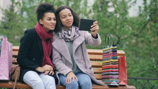 Dos mujeres afroamericanas alegres con bolsas de compras que tienen videollamada con tableta. Jóvenes amigos sentados en el banco tienen chat en línea — Vídeos de Stock