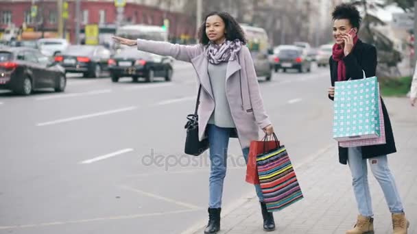 Dos atractivas mujeres afroamericanas con bolsas de compras pidiendo taxi mientras regresan de las ventas del centro comercial — Vídeos de Stock