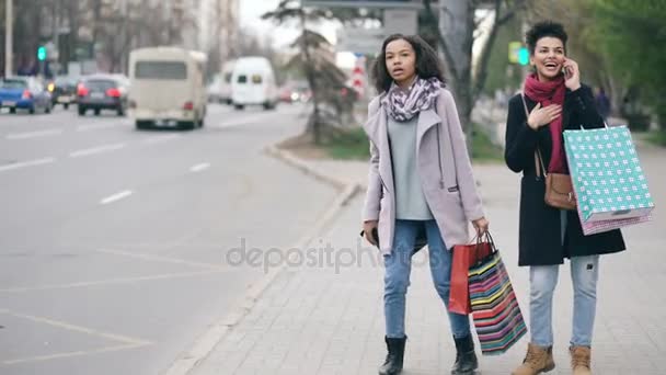 Dos atractivas mujeres afroamericanas con bolsas de compras pidiendo taxi mientras regresan de las ventas del centro comercial — Vídeos de Stock