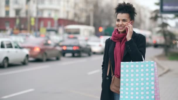 Vonzó afrikai-amerikai nő beszél a telefon rövid idő várakozás-re taxi taxi, és jön vissza a mall értékesítési bevásárló táskák — Stock videók