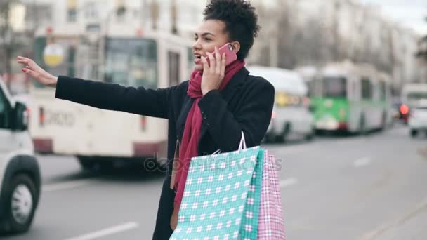 Atractiva mujer afroamericana con bolsas de compras hablando por teléfono mientras espera un taxi y regresa de las ventas del centro comercial — Vídeo de stock