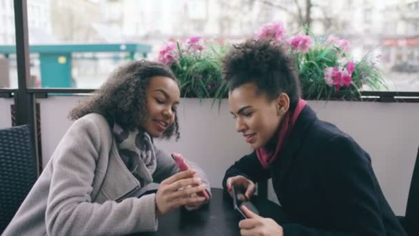 Two attractive mixed race female friends sharing together using smartphone in street cafe outdoors — Stock Video