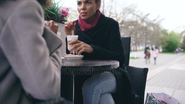 Inclinado hacia arriba de dos atractivas mujeres de raza mixta hablando y tomando café en la cafetería de la calle. Amigos se divierten después de visitar la venta del centro comercial — Vídeos de Stock