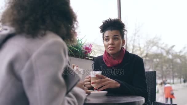 Két vonzó vegyes verseny nők beszélő és iszik kávét, utcai kávézó. Meg szórakozni után látogat a mall eladó — Stock videók