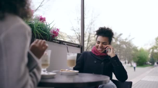 Dos mujeres de raza mixta sentadas a la mesa en la cafetería de la calle, usando teléfonos inteligentes y hablando — Vídeo de stock