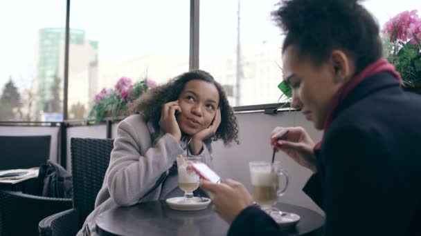 Mulher de raça mista atraente sentada à mesa no café de rua falando telefone celular enquanto seu amigo surfar smartphone — Vídeo de Stock