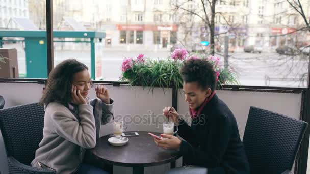 Atractiva mujer de raza mixta sentada a la mesa en la cafetería de la calle hablando de teléfono celular mientras su amigo navega por teléfono inteligente — Vídeo de stock