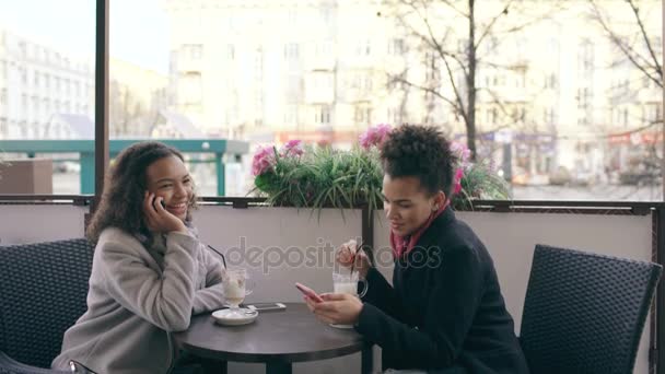Atractiva mujer de raza mixta sentada a la mesa en la cafetería de la calle hablando de teléfono celular mientras su amigo navega por teléfono inteligente — Vídeos de Stock