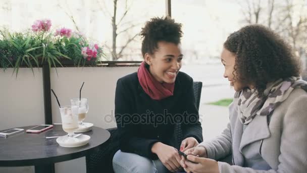 Két vonzó vegyes verseny nők beszélő és iszik kávét, utcai kávézó. Meg szórakozni után látogat a mall eladó — Stock videók