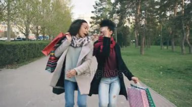İki çekici karışık yarış bayanlar dans çekim dolly ve eğlenceli alışveriş torbaları ile park aşağı yürürken. Alışveriş Merkezi satış ziyaret ettikten sonra mutlu genç arkadaş yürüyüş