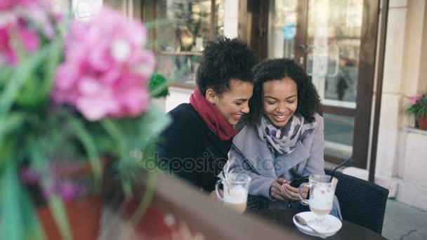 Dos atractivas amigas de raza mixta compartiendo juntos el uso de smartphone en la cafetería al aire libre — Vídeos de Stock