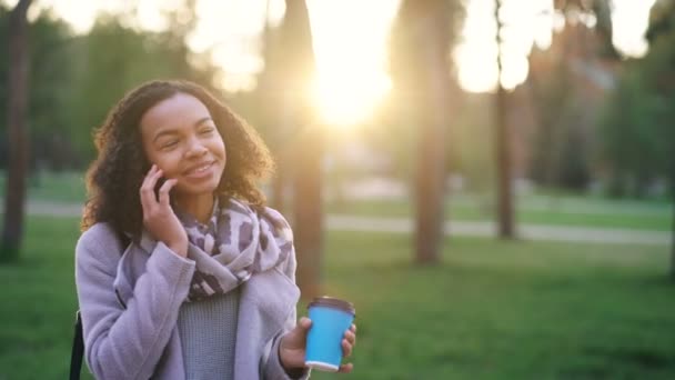 Aantrekkelijke gemengd ras meisje praten smartphone en drinken koffie in stadspark met zakken. Jonge vrouw na het winkelen winkelcentrum uitverkoop — Stockvideo