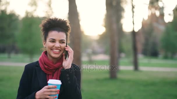 Aantrekkelijke gemengd ras meisje praten smartphone en drinken koffie in stadspark met zakken. Jonge vrouw na het winkelen winkelcentrum uitverkoop — Stockvideo