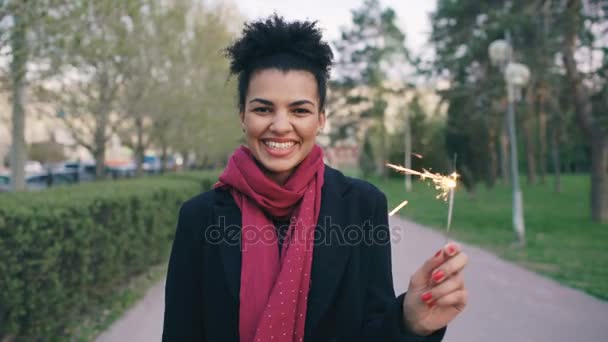 Attraente razza mista giovane donna accende un fuoco d'artificio scintillante e sorridente nel parco — Video Stock