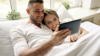 Tablet bilgisayar kullanarak ve yatak odasında yatakta sabah yalan konuşuyor genç tatlı ve sevgi dolu Çift