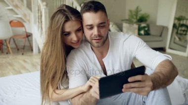 Tatlı ve sevgi dolu bir genç çift tablet bilgisayar kullanarak, sabah yatakta yatak odasında otururken öp