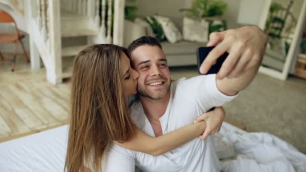 Fechar-se de jovem casal bonito e amoroso tirar foto selfie na câmera do smartphone e beijar enquanto sentado na cama na manhã — Vídeo de Stock