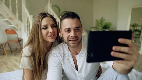 Joven linda y cariñosa pareja de tener video chat celebración de la tableta de ordenador y charlar con amigos sentados en la cama en casa — Vídeo de stock