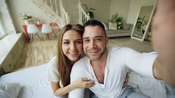 Jovem casal bonito e amoroso ter vídeo chat segurando smartphone e conversando com os pais sentados na cama em casa — Vídeo de Stock