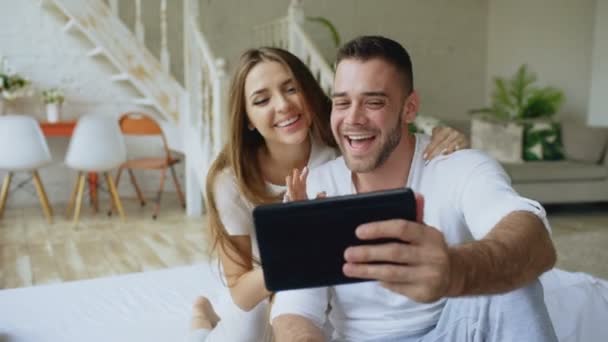 Joven hombre atractivo utilizando la tableta de ordenador que tiene chat de vídeo, mientras que su novia vienen y lo abrazan. Linda pareja amorosa charlando con los padres sentados en la cama en casa — Vídeo de stock