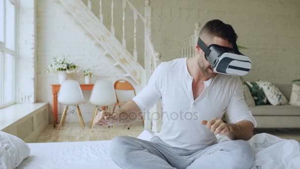 Χαρούμενος νεαρός φορώντας σετ κεφαλής εικονικής πραγματικότητας που παίζει το παιχνίδι βίντεο 360 Vr ενώ κάθεται στο κρεβάτι στο σπίτι — Αρχείο Βίντεο