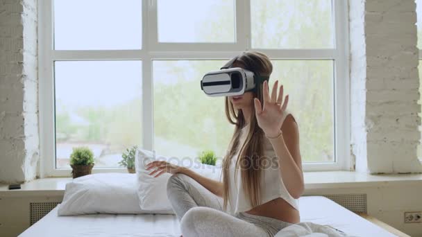 Молодая веселая женщина в наушниках виртуальной реальности, играющая в видеоигру 360 VR, сидит дома в постели — стоковое видео