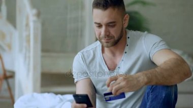 Çekici genç adam akıllı telefon ve internet oturup evde yatakta alışveriş kredi kartı ile