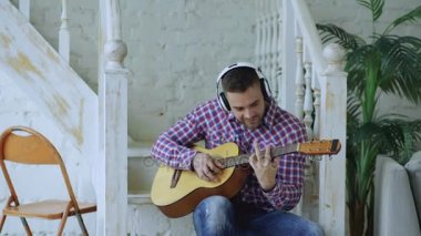 Kablosuz kulaklık ile merdivenlerde oturan mutlu çekici adam oyun akustik gitar evde çalışma