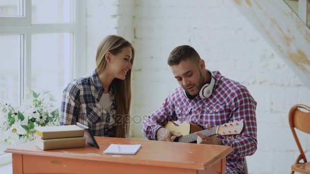 Pasangan muda yang bahagia dan penuh kasih belajar untuk bermain gitar akustik dan bersenang-senang sambil duduk di meja di rumah — Stok Video