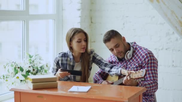 Joven pareja feliz y cariñosa estudio para tocar la guitarra acústica utilizando tableta y divertirse mientras se sienta en la mesa en casa — Vídeos de Stock