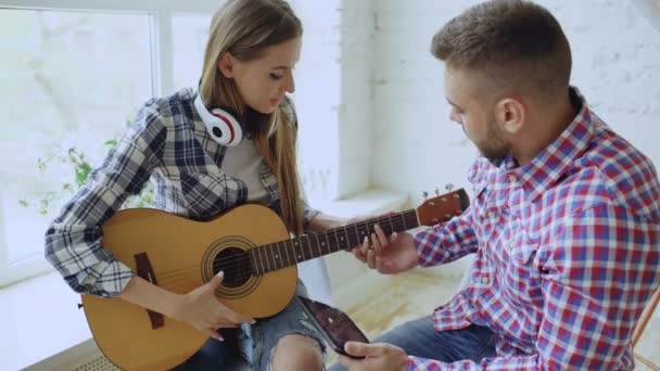 Молодая счастливая и любящая пара учится играть на акустической гитаре с помощью планшетного компьютера и веселиться, сидя за столом дома — стоковое видео