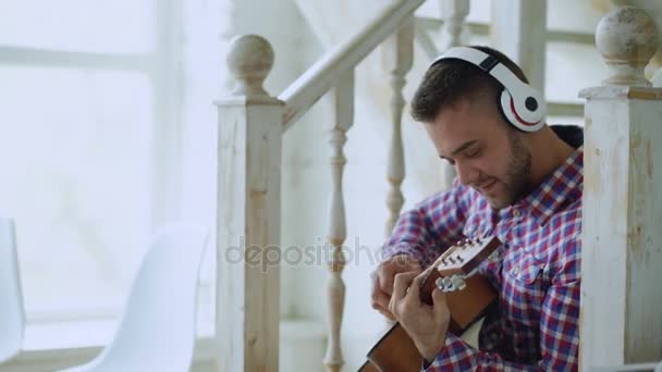 Lycklig attraktiv man sitter på trappan med trådlösa hörlurar studera spela akustisk gitarr hemma — Stockvideo