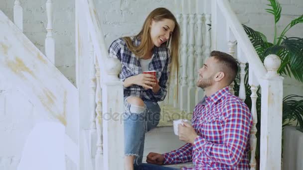 Молодая счастливая и любящая пара пьет чай и разговаривает, сидя на лестнице в гостиной на дому — стоковое видео