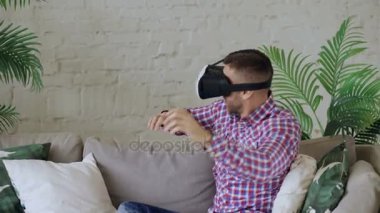 Evde oturma odasında kanepede otururken 360 Vr video oyun oynarken giyiyor sanal gerçeklik kulaklık neşeli genç adam