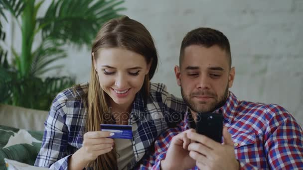 スマート フォンと自宅のリビング ルームでソファの上にインターネット座るでショッピング クレジット カードと幸せな若いカップルのクローズ アップ — ストック動画